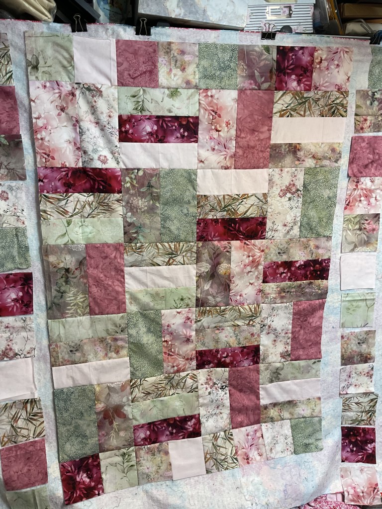 A patchwork quilt top featuring a mix of floral and geometric patterned fabric pieces in shades of pink, green, and cream, arranged in a grid layout.