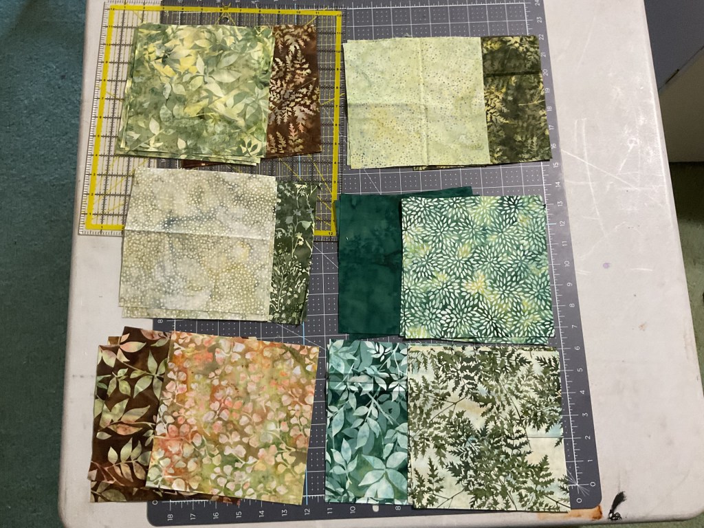 A variety of fabric squares in shades of green and brown, arranged on a cutting mat, ready for a quilting project.