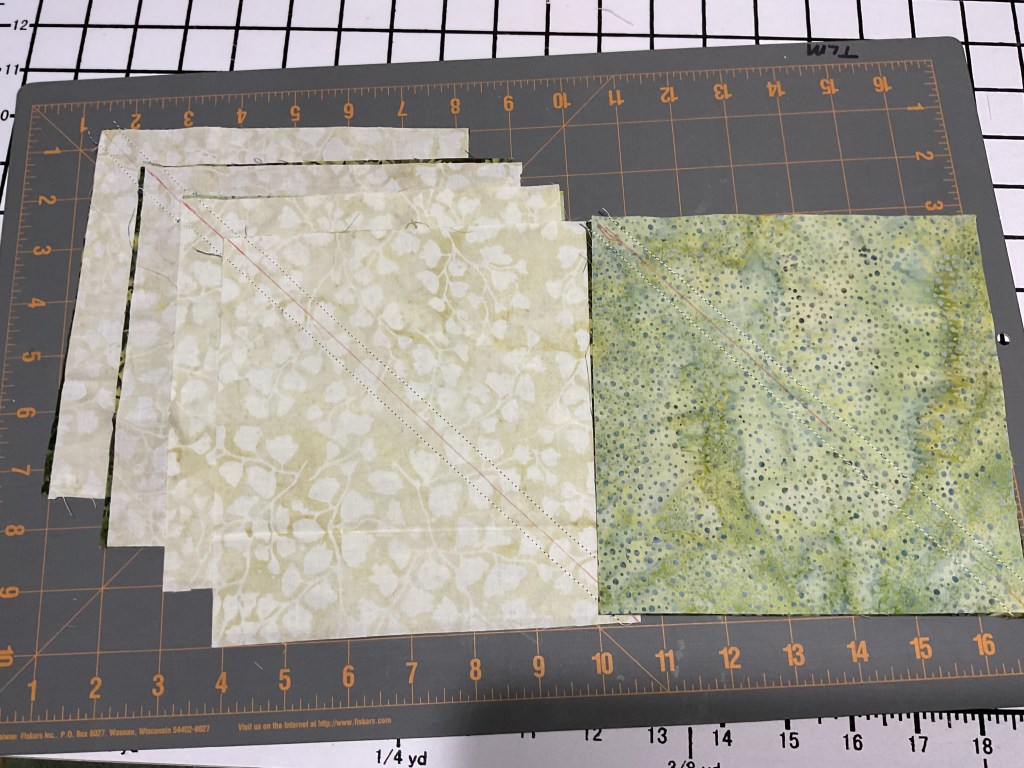 A selection of cut fabric pieces arranged on a rotary cutting mat, with a marked diagonal line on one piece, ready for sewing.