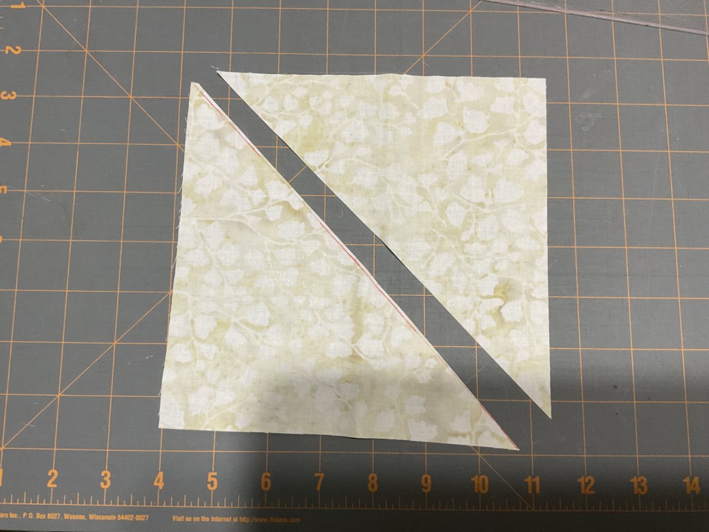A diagonal cut through a light green fabric square with a floral pattern, placed on a cutting mat with grid lines.