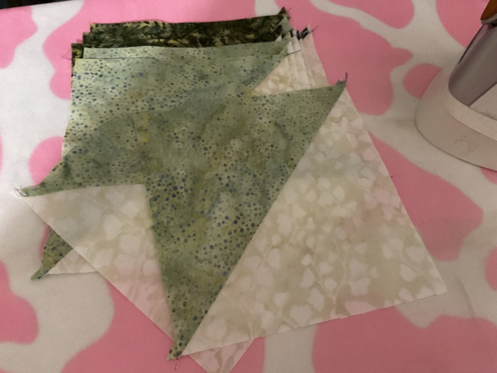 A selection of cut quilt fabric pieces in various shades of green and beige, arranged on a soft pink background.