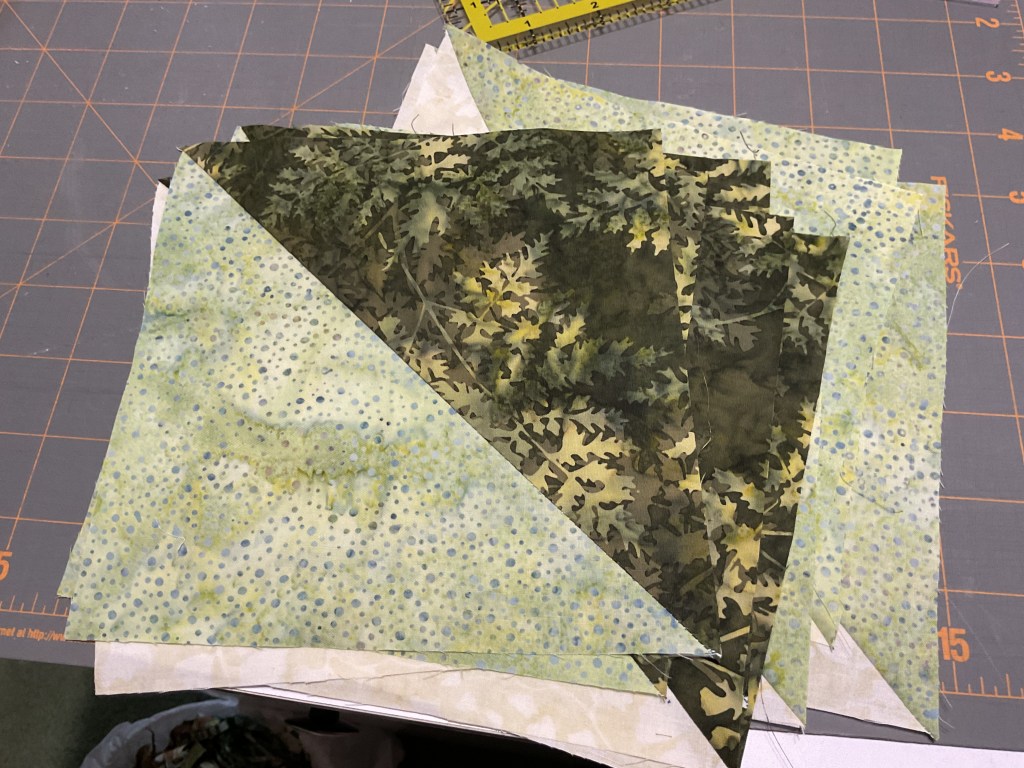 A collection of fabric pieces in shades of green and beige, cut for a quilt project, displayed on a cutting mat.