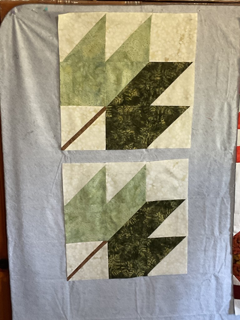 Two quilt blocks featuring stylized green and brown leaves on a light background, displayed on a blue fabric surface.