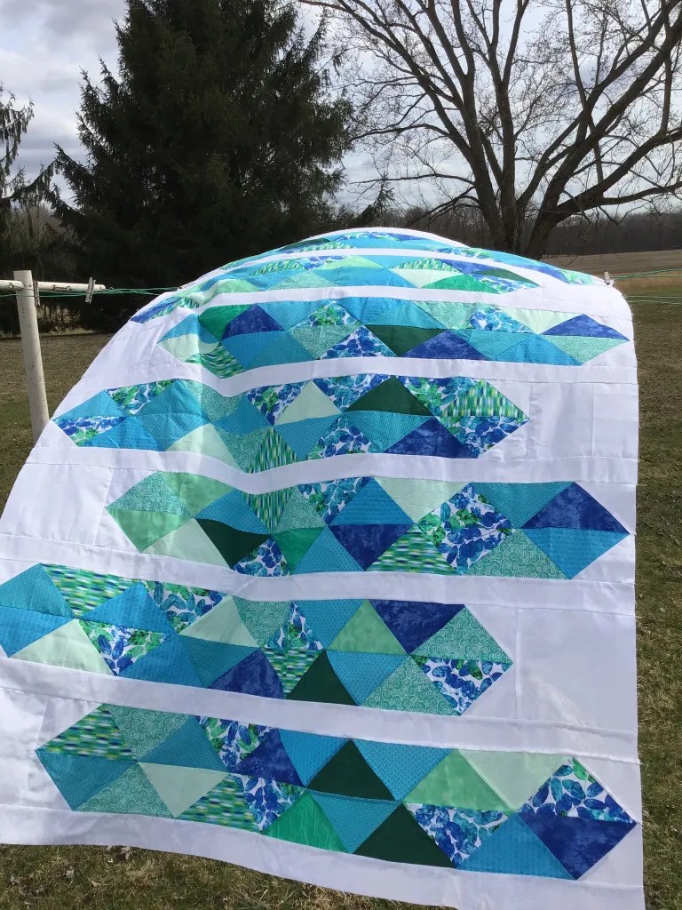 A completed quilt with geometric patterns in shades of blue and green, displayed outdoors against a backdrop of trees and a cloudy sky.