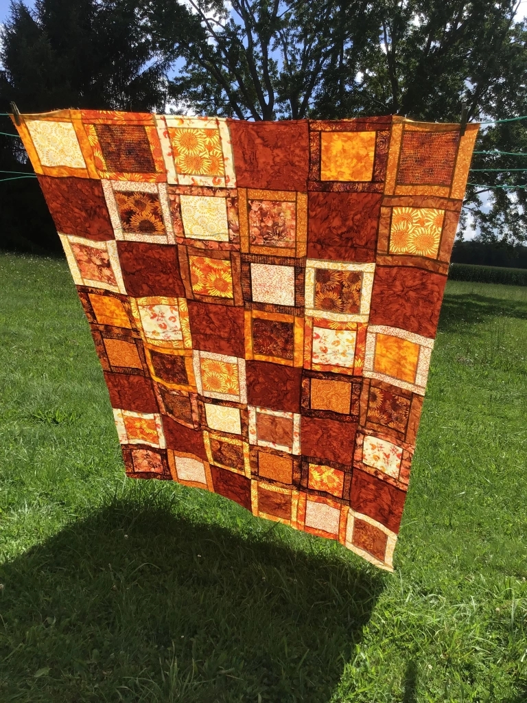 A vibrant quilt featuring warm autumn colors such as orange and brown, displayed outdoors on a sunny day, with trees and grass in the background.