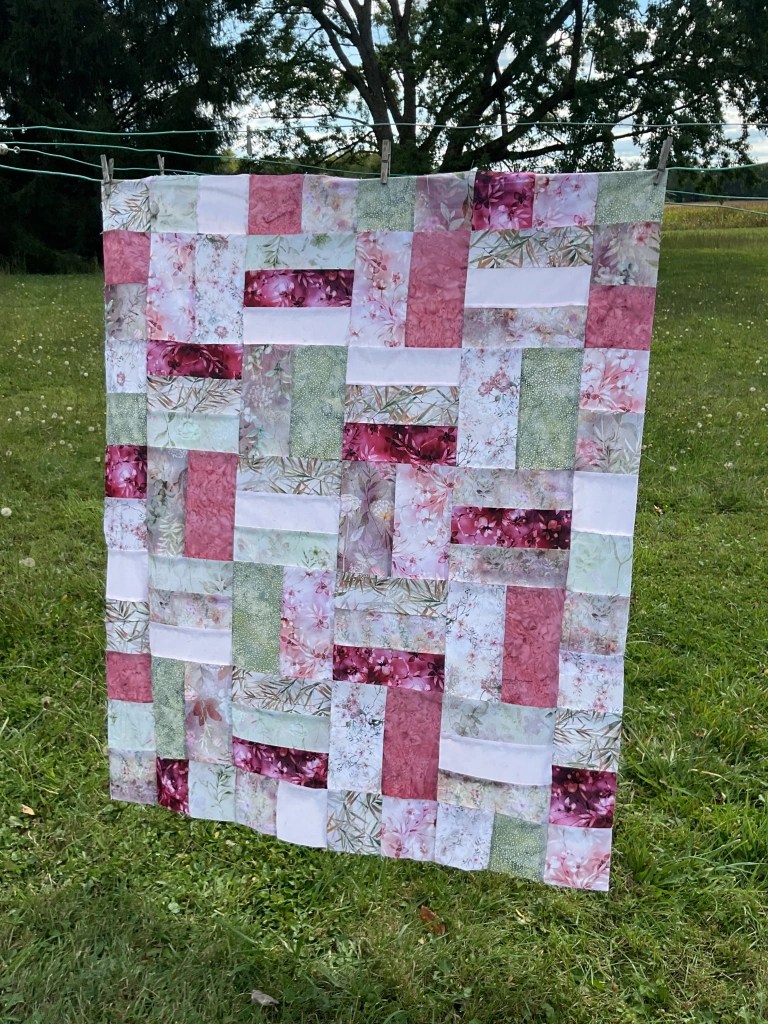 A patchwork quilt hanging on a clothesline, featuring a mix of floral and patterned fabric squares in shades of pink, green, and white.