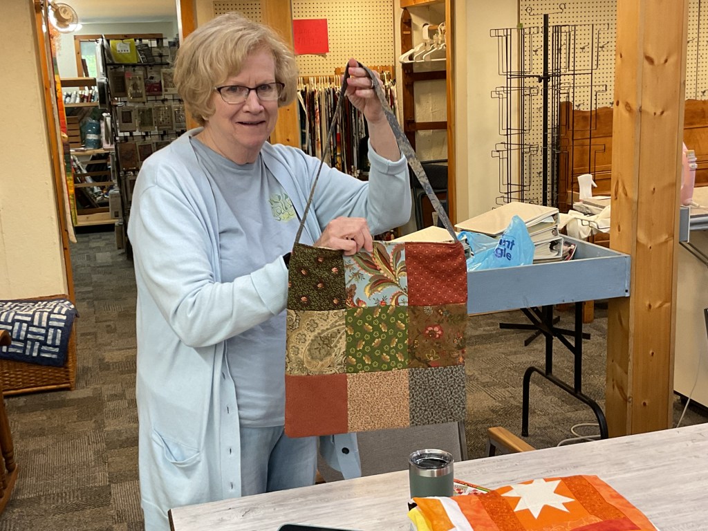 A woman holding a handcrafted patchwork bag, displaying various fabric patterns and colors, in a sewing space filled with quilting materials and supplies.