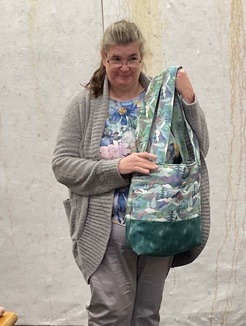 A woman wearing a floral shirt and a gray cardigan is holding a colorful tote bag with a scenic design, smiling at the camera.