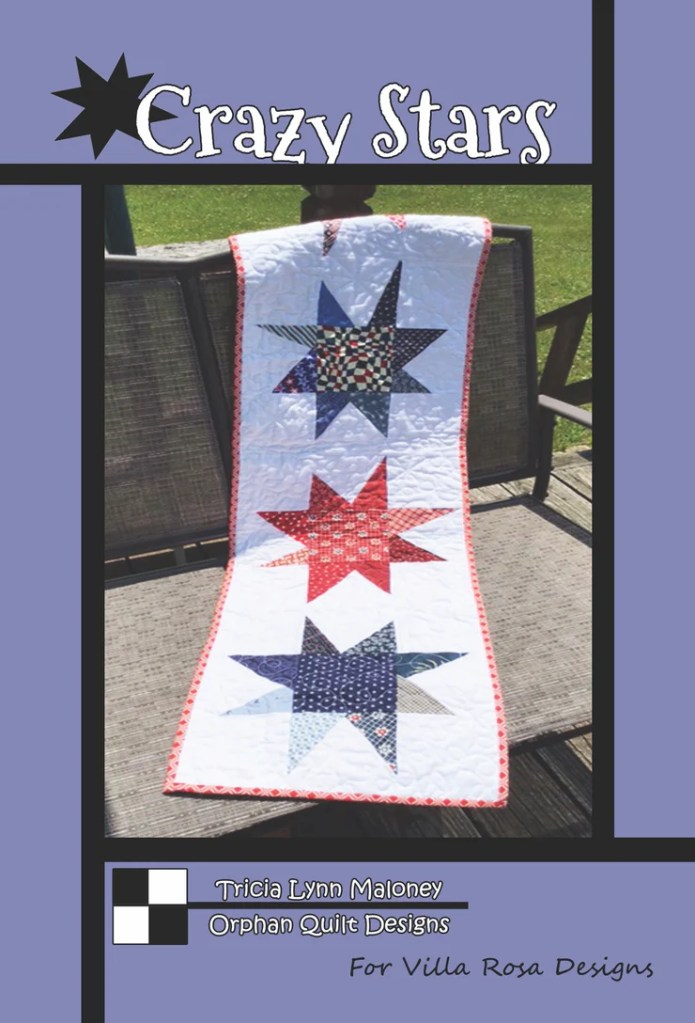 Cover image of the Crazy Stars quilt pattern featuring a table runner with colorful star designs in red, blue, and white. The quilt is displayed on a wooden bench against a green background.