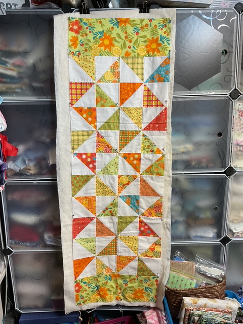 A completed table runner featuring a pinwheel design in vibrant colors including orange, yellow, green, and floral patterns, hanging against a backdrop of organized fabric shelves.
