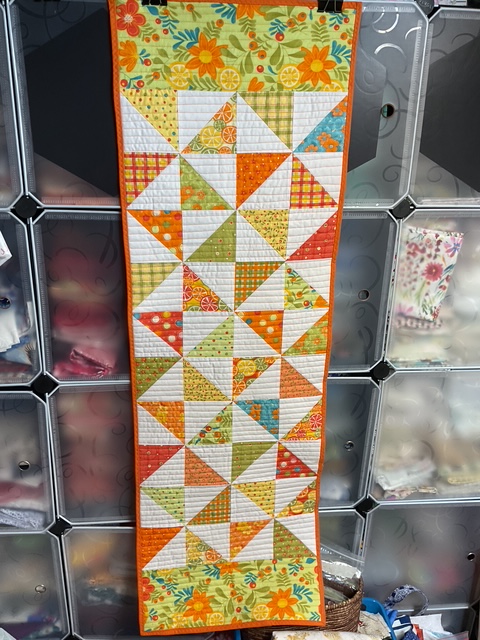 A vibrant quilted table runner featuring a pinwheel pattern in colorful fabrics, including orange, green, and yellow, displayed against a backdrop of fabric storage.
