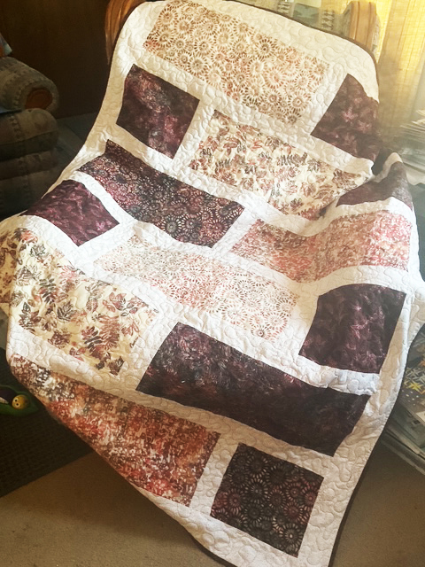 A quilt with a patchwork design featuring rich burgundy, cream, and floral prints, displayed draped over a chair in a cozy setting.