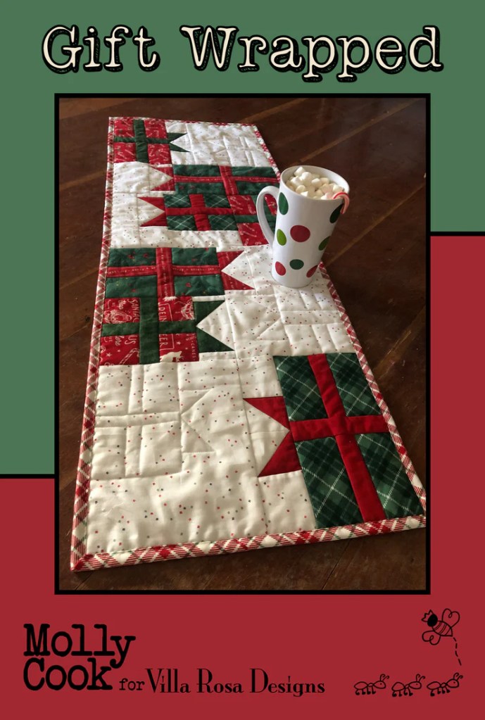 A holiday-themed quilted table runner titled 'Gift Wrapped' featuring red, green, and white patterns, accompanied by a polka dot mug filled with marshmallows.