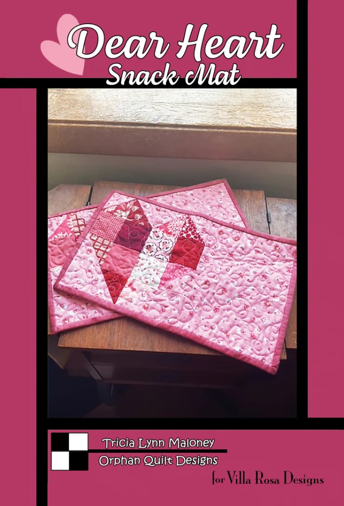 A pink quilted snack mat featuring a heart design, with the text 'Dear Heart Snack Mat' prominently displayed. The mat is placed on a wooden surface near a window.
