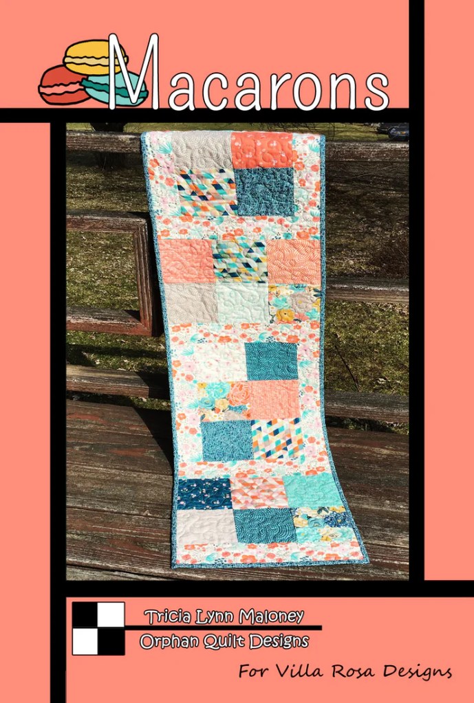 A quilted table runner featuring a colorful patchwork design with aqua, coral, and floral patterns, titled 'Macarons' by Tricia Lynn Maloney, set against a warm pink background.