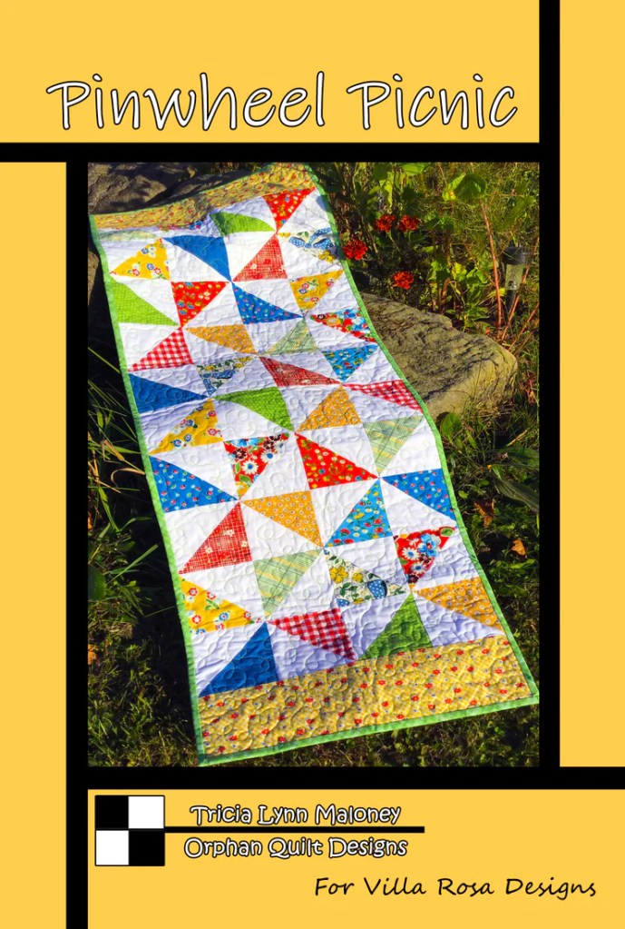 A vibrant quilted table runner called 'Pinwheel Picnic' features colorful triangular patterns in various fabrics, set against a sunny yellow background. The runner showcases a mix of floral and checkered designs, emphasizing a playful summer theme.