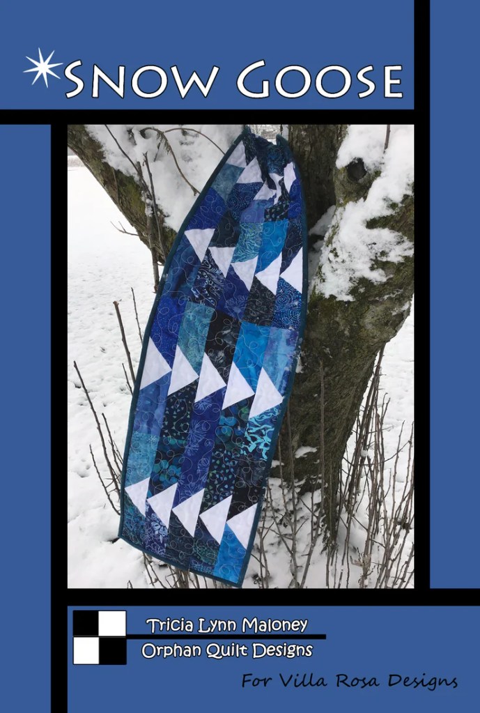 A quilt titled 'Snow Goose' featuring blue and white geometric patterns, hanging on a tree branch amidst a snowy background.