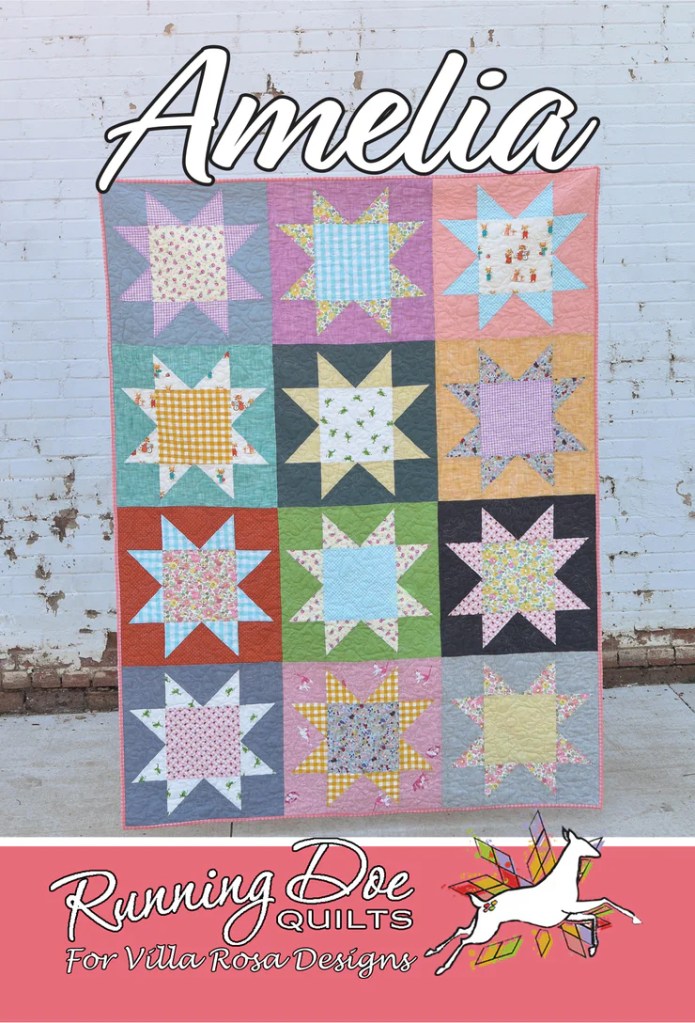 A colorful quilt titled 'Amelia' featuring star patterns in various fabrics, displayed against a white brick background.