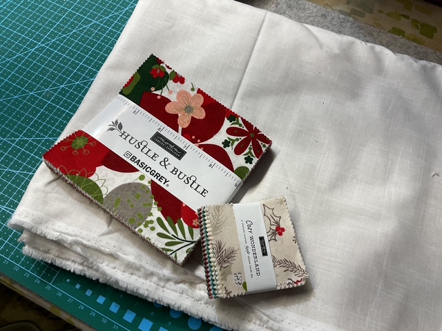 A stack of fabric pieces including a charm pack with holiday patterns and a white fabric, placed on a green cutting mat.