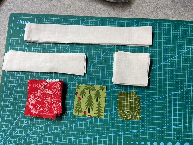 A variety of fabric pieces laid out on a cutting mat, including red patterned squares, green squares with pine tree designs, and strips of white fabric.