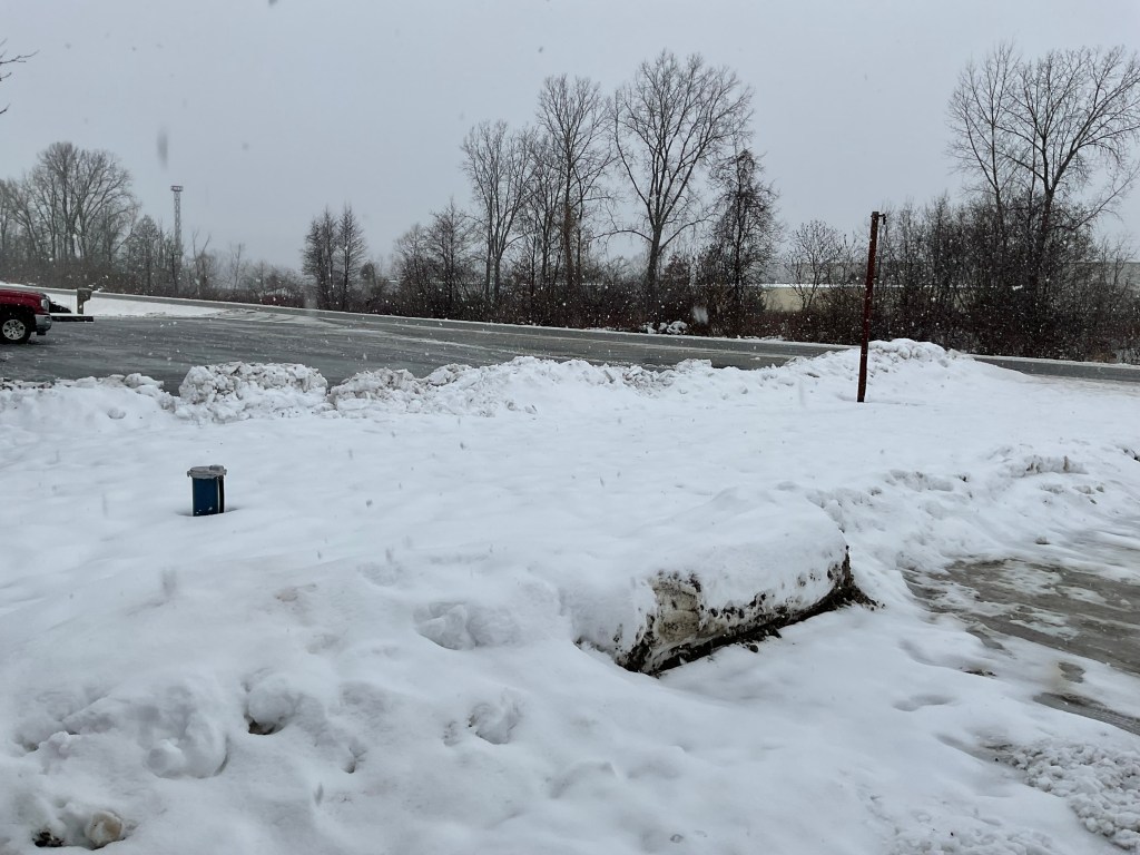 A snowy winter scene with a parking lot covered in snow and slush. In the background, trees are bare, and snow is falling gently from the gray sky.