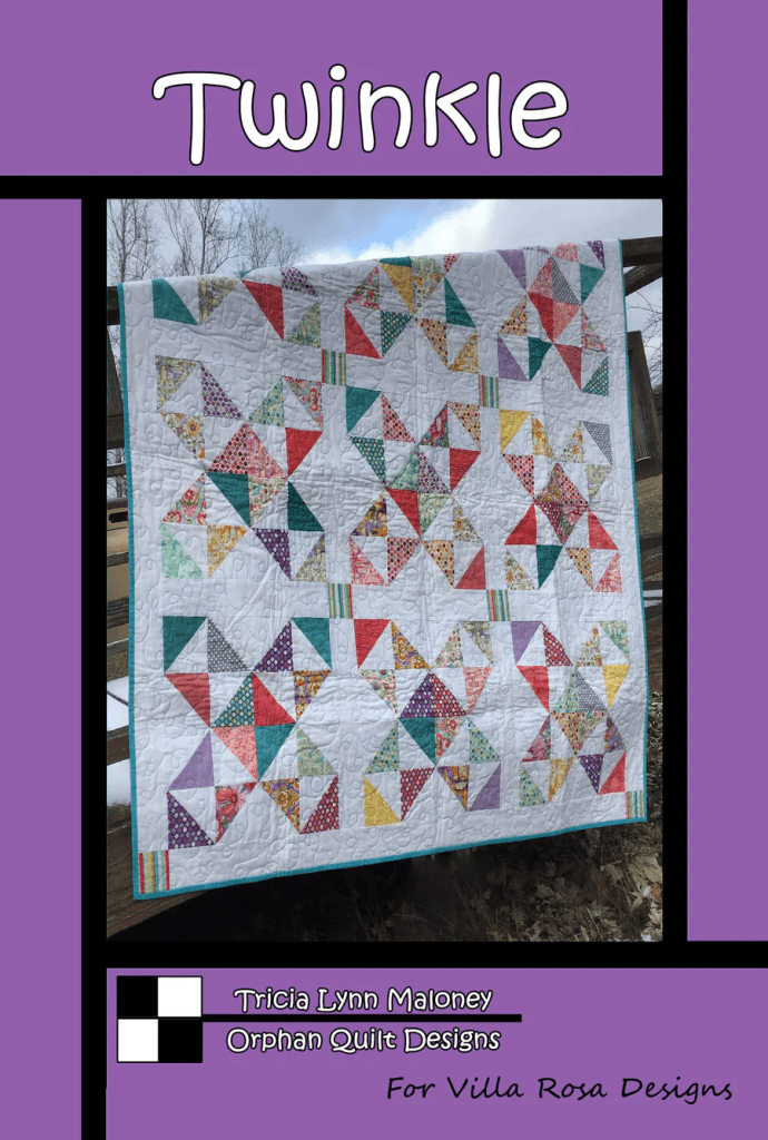 A colorful quilt titled 'Twinkle' featuring a geometric pattern of triangles in various fabrics hanging outdoors.