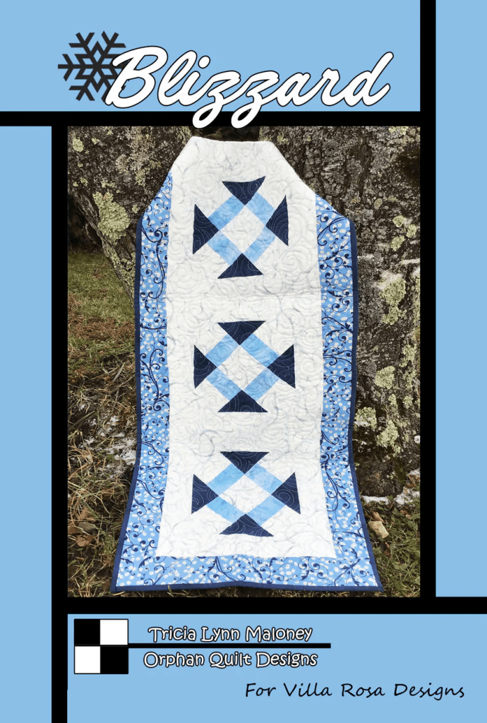 A blue and white quilt displayed outdoors against a tree, featuring a snowflake design and bordered by floral patterns. Text at the top reads 'Blizzard' and includes designer credits.