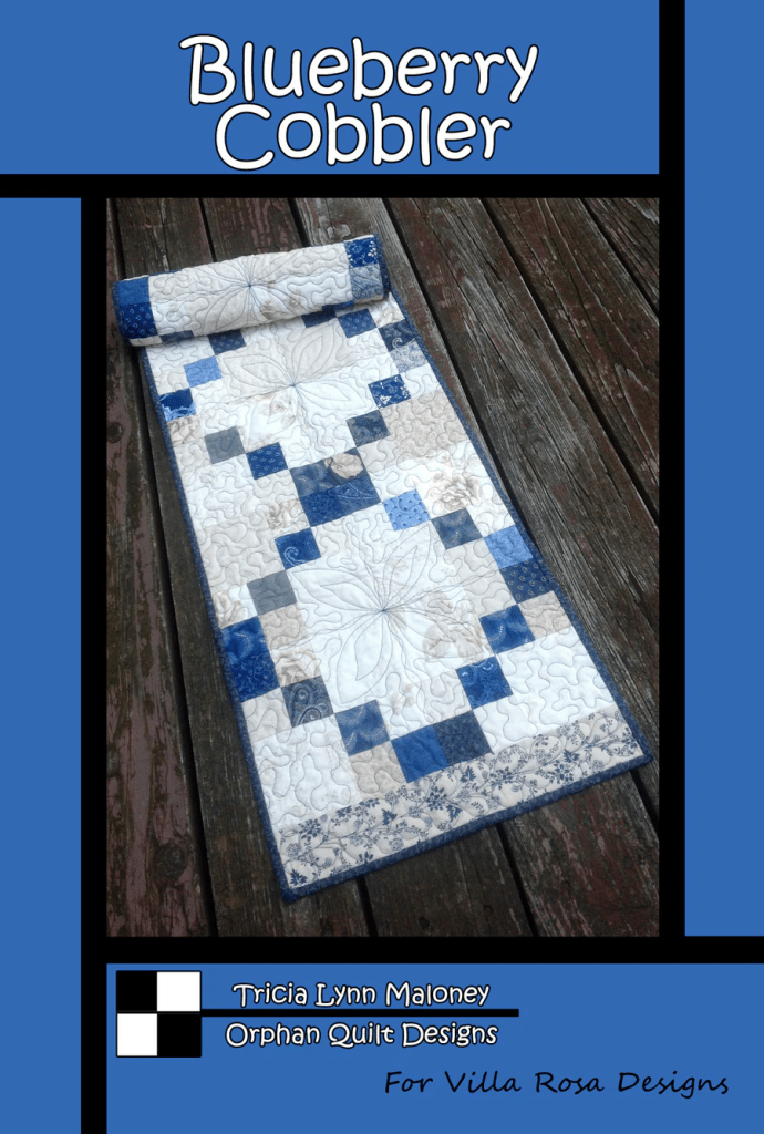 A quilt titled 'Blueberry Cobbler' by Tricia Lynn Maloney, featuring a patchwork design in shades of blue, cream, and beige, displayed on a wooden surface.