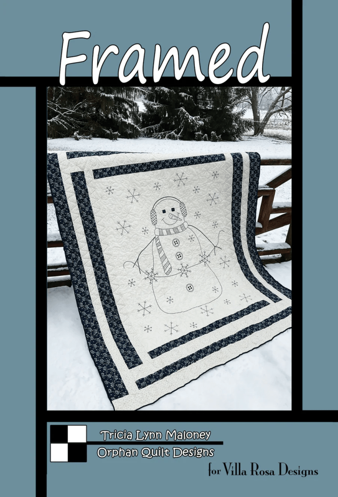 A quilt featuring a snowman design outlined in black against a white background, bordered by navy fabric with a snowflake pattern, displayed outdoors in a snowy setting.