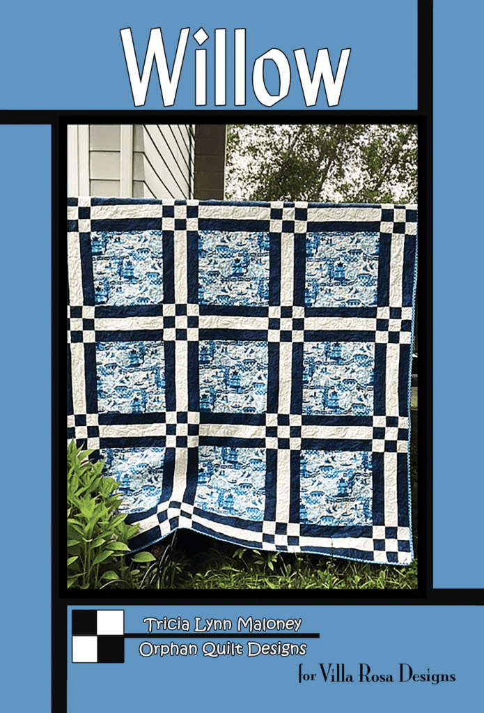 A blue-themed quilt featuring a checkered pattern with white and navy blocks, displayed outdoors against a light-colored wall, with greenery in the foreground.