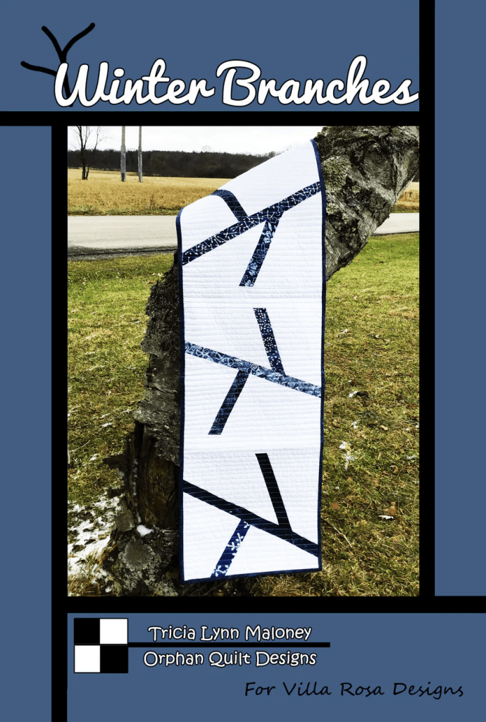 A quilt named 'Winter Branches' hanging on a tree stump, featuring a modern design with blue and white branches on a neutral background, against a rural landscape.