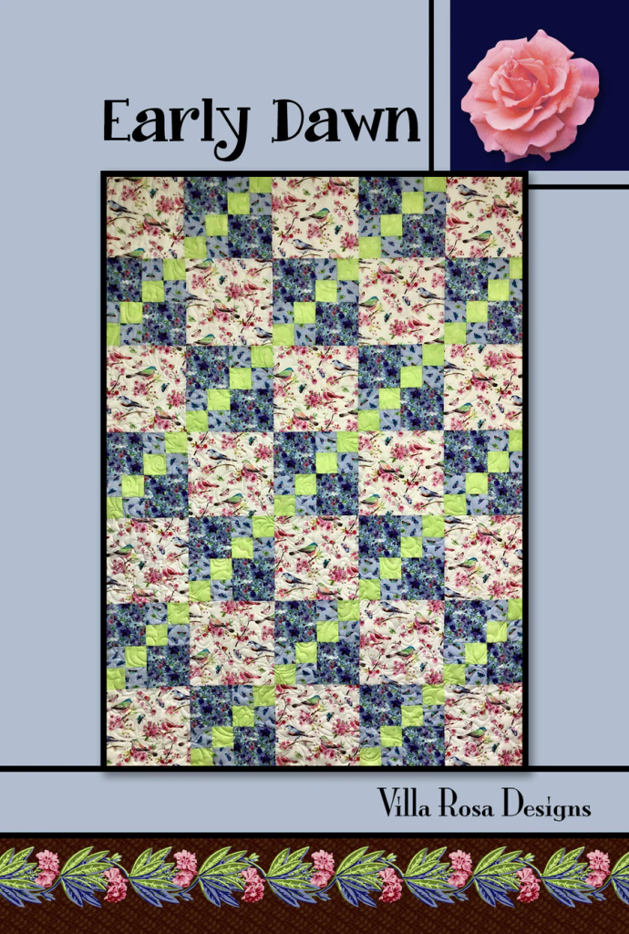A colorful quilt titled 'Early Dawn' by Villa Rosa Designs, featuring a patchwork pattern of floral prints in pink, blue, green, and cream. The quilt is framed with a decorative floral border.