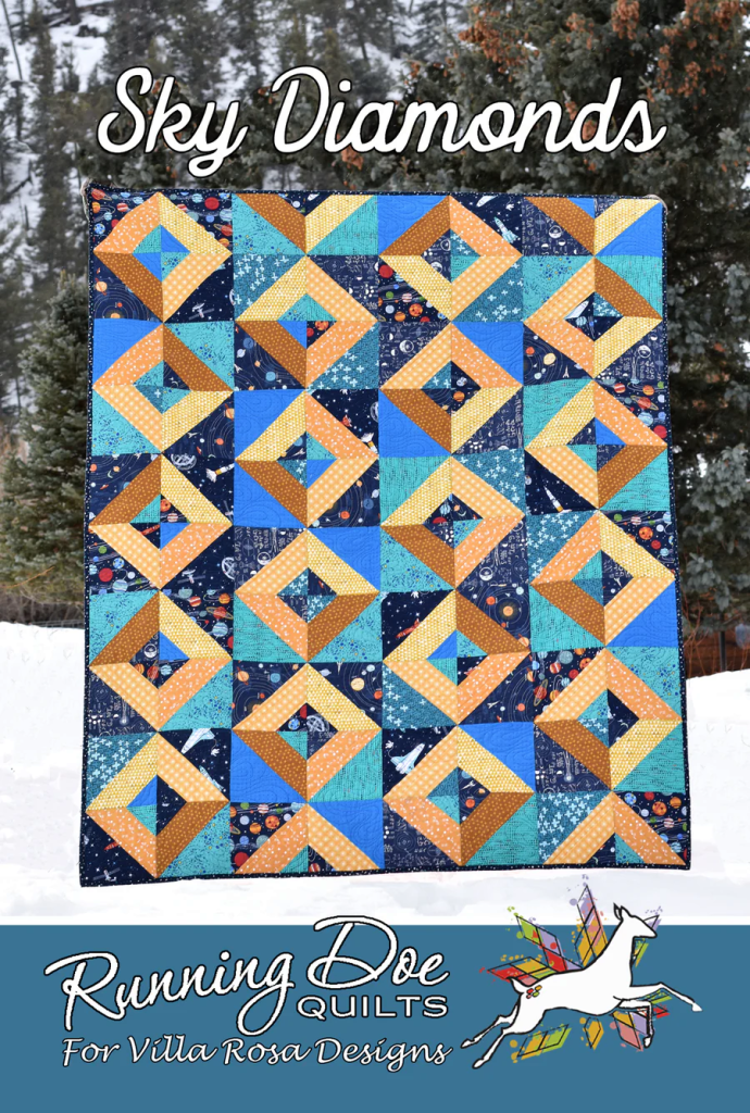 A vibrant quilt titled 'Sky Diamonds' featuring geometric patterns in shades of blue, orange, and yellow, displayed against a snowy outdoor backdrop.