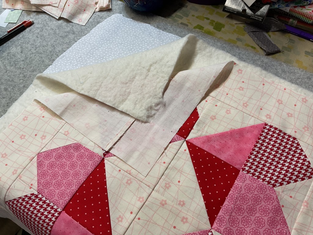 A close-up view of a quilting project showing a variety of pink and red fabric squares arranged in a geometric pattern, with batting visible underneath.