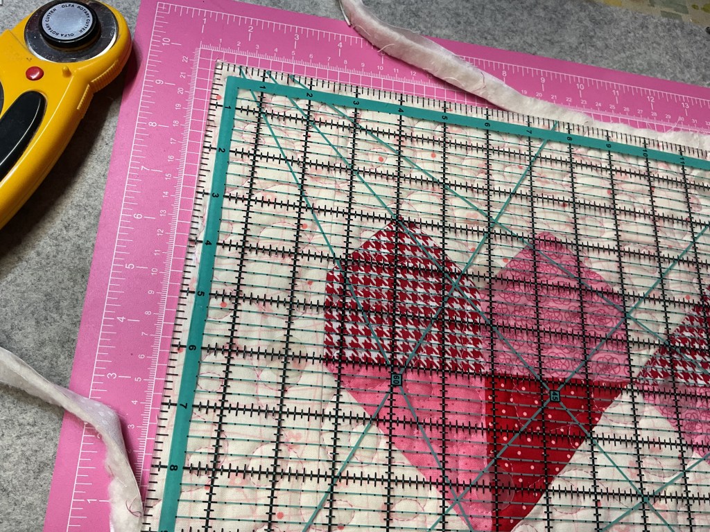 A cutting mat with a transparent ruler and rotary cutter, featuring a patchwork design of hearts in red and pink fabrics.