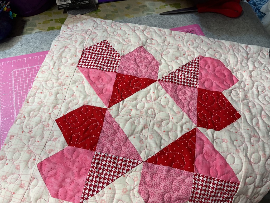 Close-up of a quilt featuring a heart pattern with red and pink fabric pieces, set against a light background with quilting stitches.
