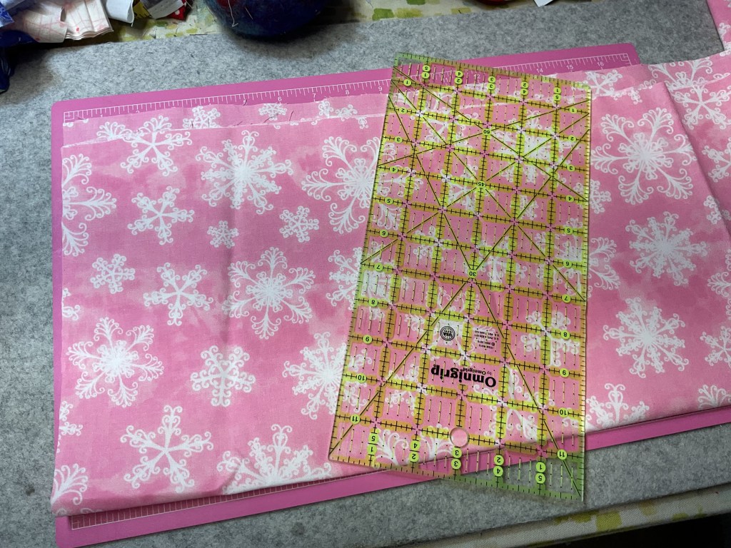 A cutting mat with pink fabric featuring white snowflakes, alongside a transparent quilting ruler marked with measurements.