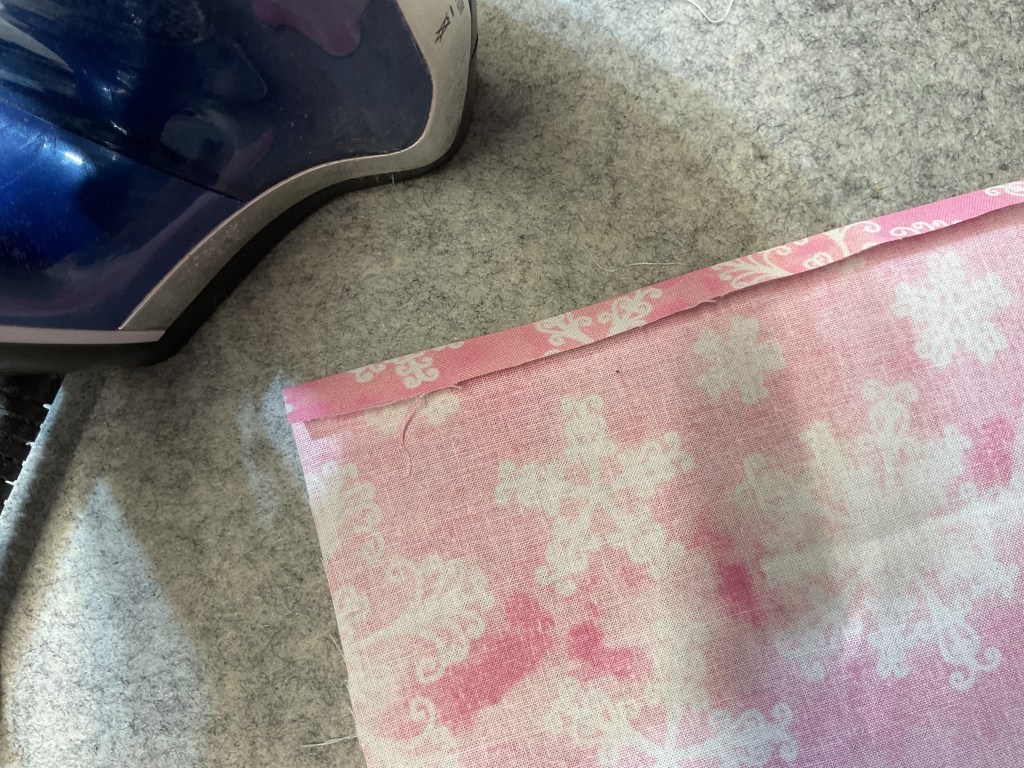 An iron next to a piece of pink fabric with a snowflake pattern, resting on a grey ironing board cover.