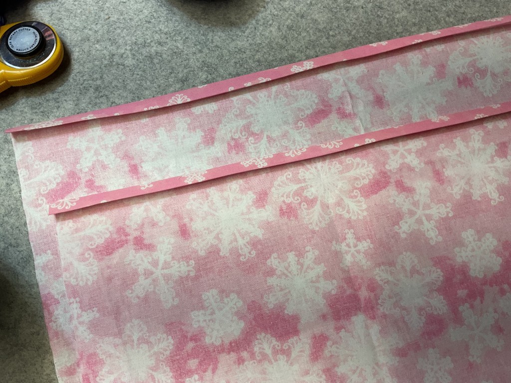 A piece of fabric featuring a pink and white snowflake pattern, with pink bias tape laid on top, on a textured surface.
