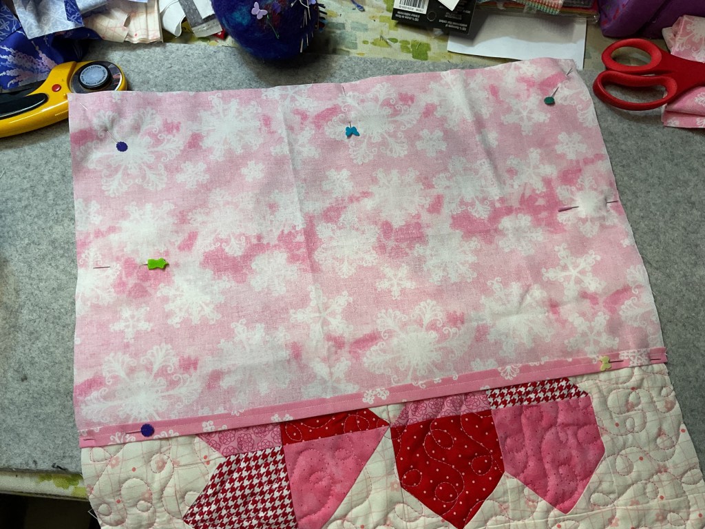 A piece of pink fabric with snowflake patterns is pinned to a quilt, surrounded by sewing tools and materials.