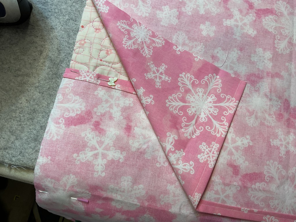 A close-up of pink fabric with a white snowflake pattern, folded and pinned for sewing.