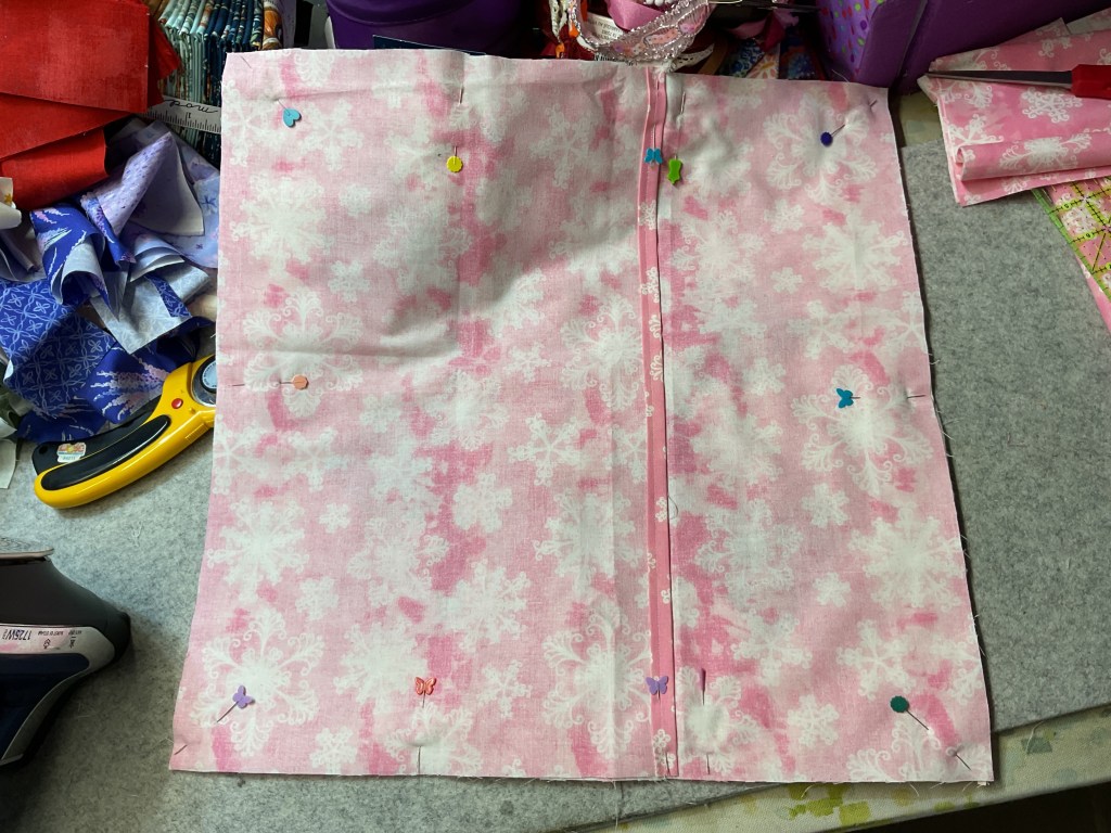 A square piece of pink fabric with a floral pattern, pinned down on a workspace with various craft supplies in the background.