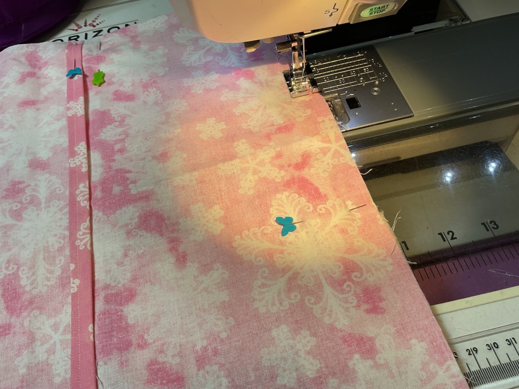 A sewing machine with a pink fabric piece featuring a floral pattern, pinned with colorful clips, under bright lighting.