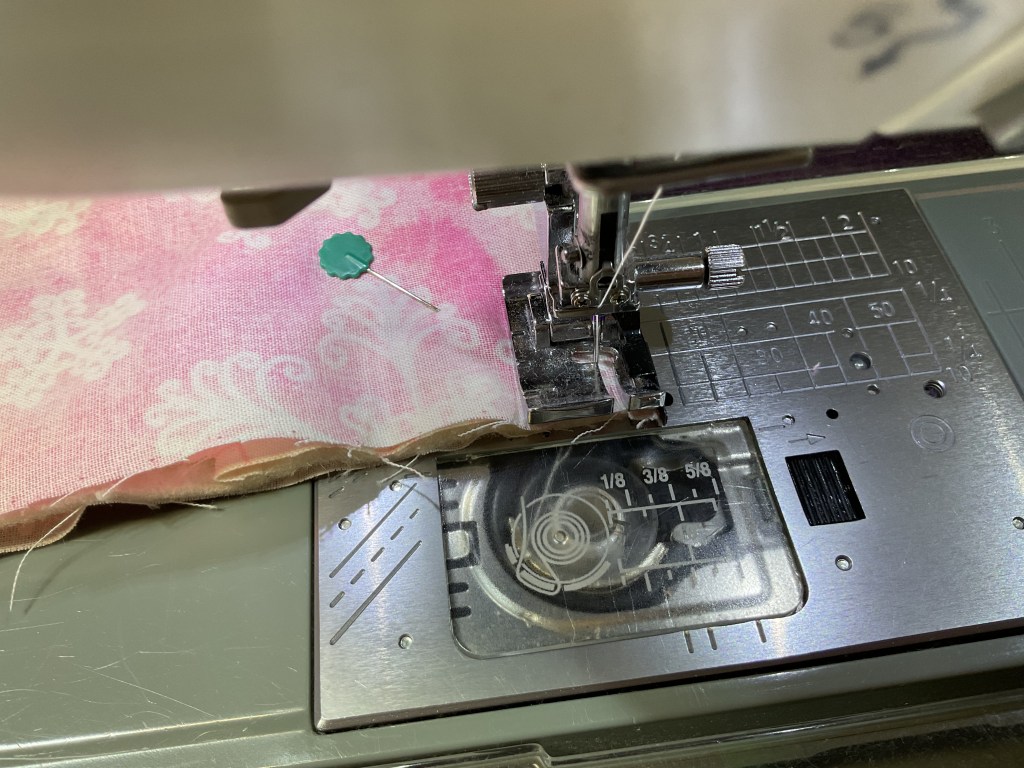 Close-up of a sewing machine foot, with pink patterned fabric being prepared for stitching, and a pin holding the fabric in place.