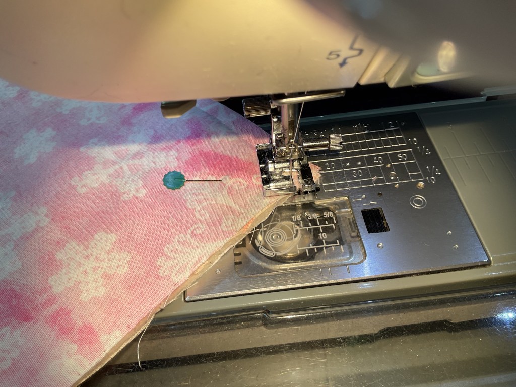 Close-up of a sewing machine with pink fabric featuring snowflake patterns and a blue sewing pin, showing the needle and presser foot in action.
