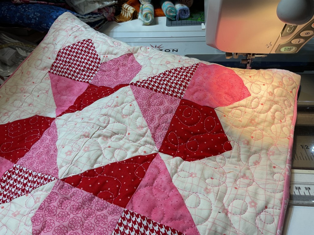 Close-up of a quilt featuring a pink and red pattern. The quilt is partially on a sewing machine with quilting lines visible, and colorful fabrics can be seen in the background.