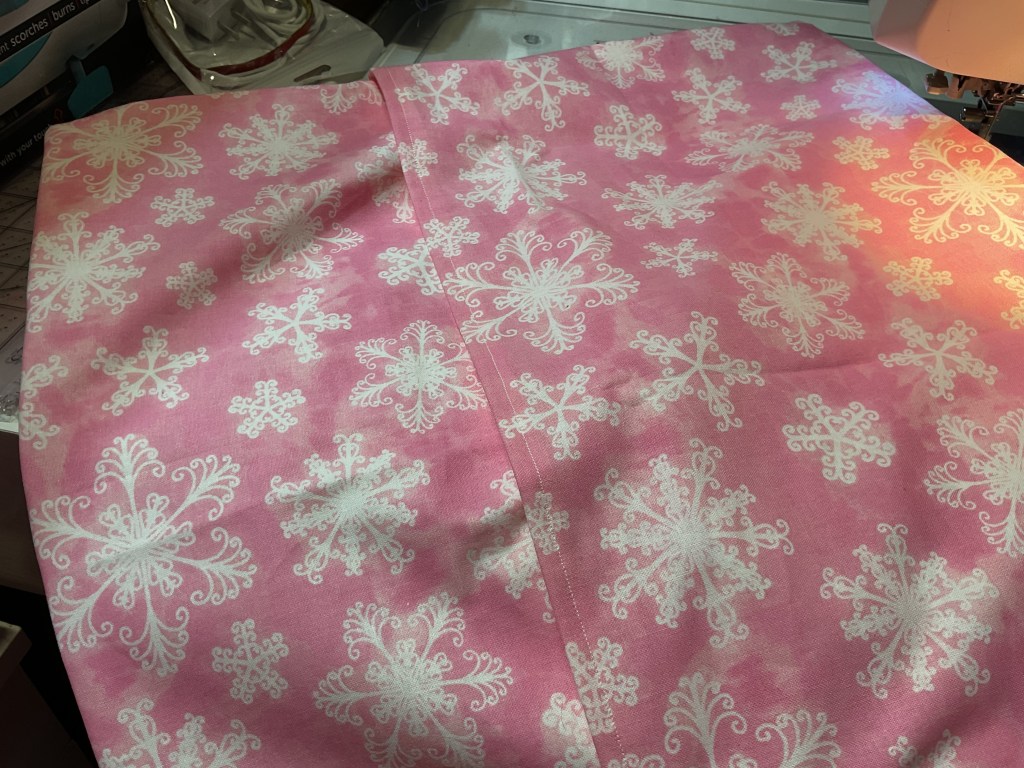 A piece of pink fabric with white snowflake patterns, laid out flat on a sewing table.