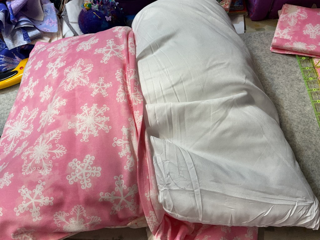 A pink pillowcase with a snowflake pattern next to a white pillow on a craft table.