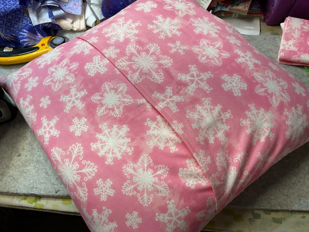 A pink pillow with white snowflake patterns, resting on a crafting table surrounded by fabric scraps and sewing tools.