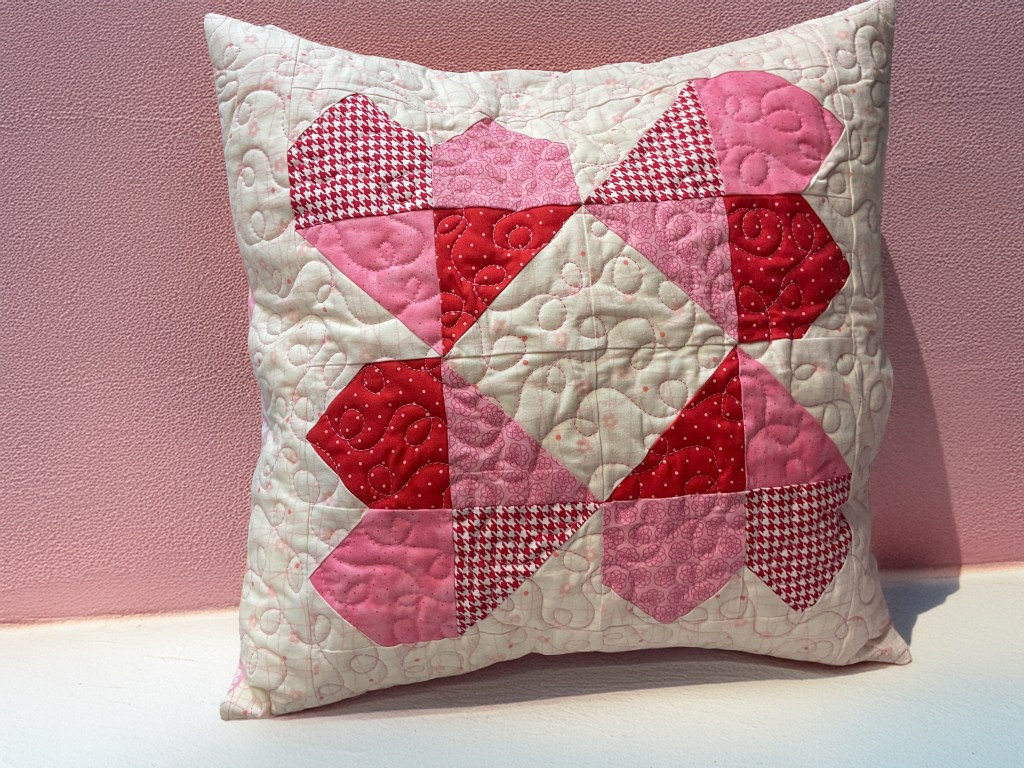 A decorative quilted pillow featuring a heart pattern in shades of pink and red against a light background.